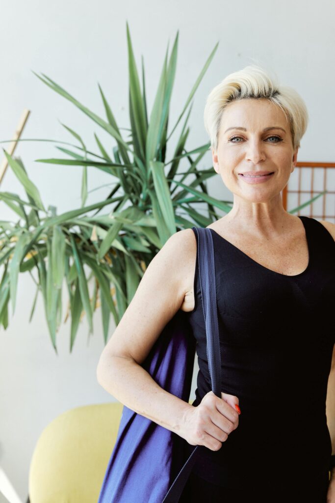 A senior woman in a black tank top smiling at the camera with a plant in the background.